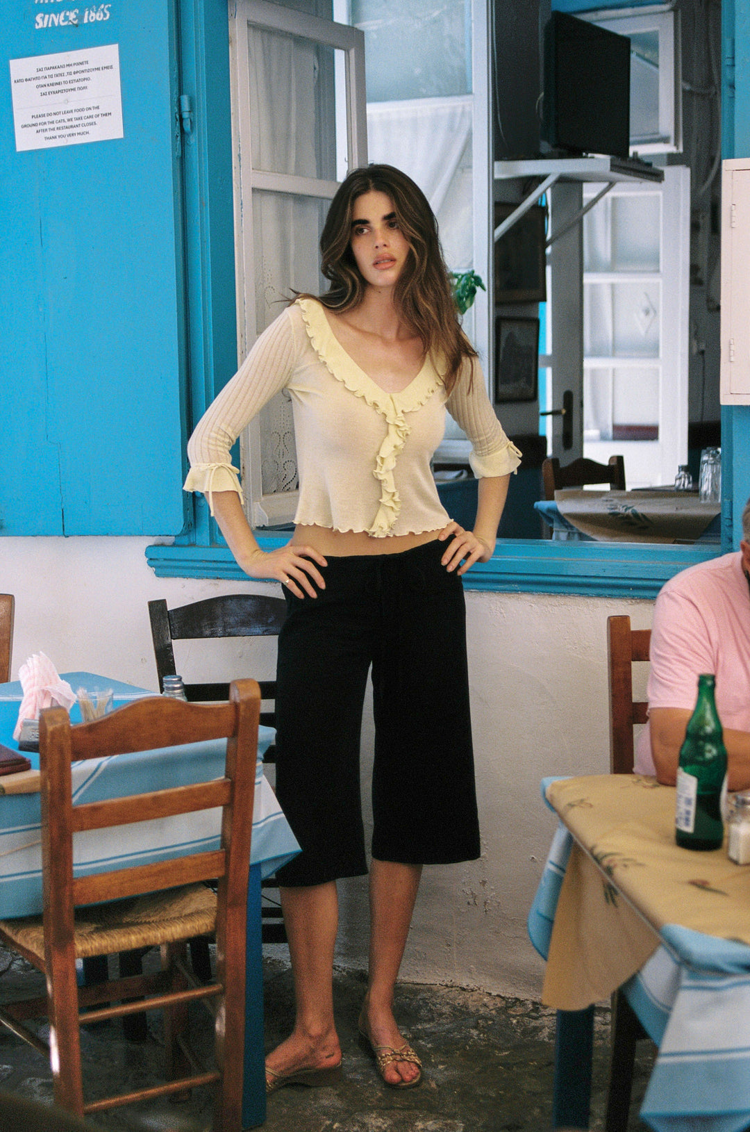 Pale yellow semi-sheer knitted top with 3/4 sleeves, ruffle-trim neckline, and tie-detail cuffs worn with black cropped pants.