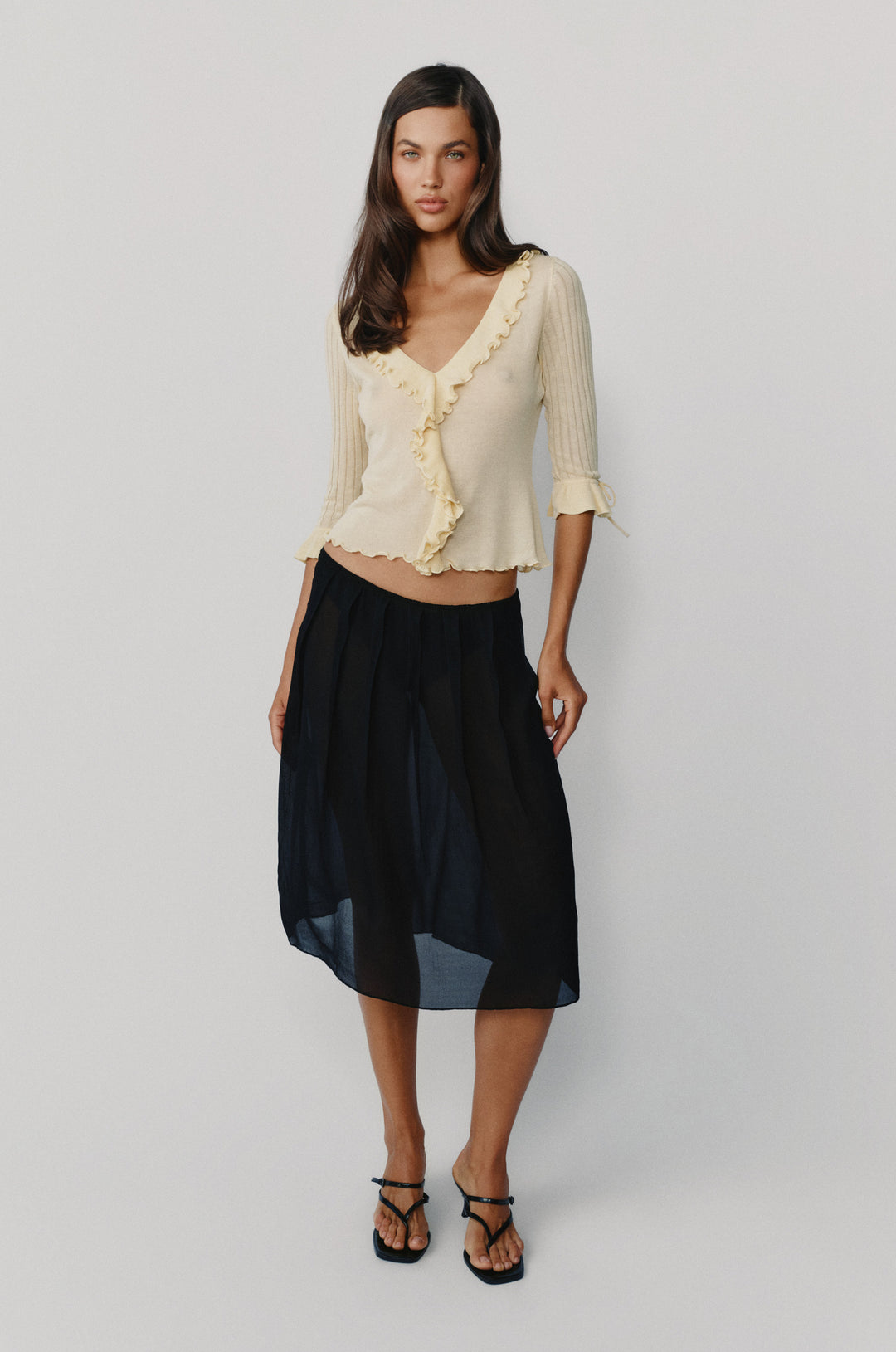 Pale yellow semi-sheer knitted top with ruffle-trim neckline, 3/4 sleeves, and tie-detail cuffs paired with a black skirt.