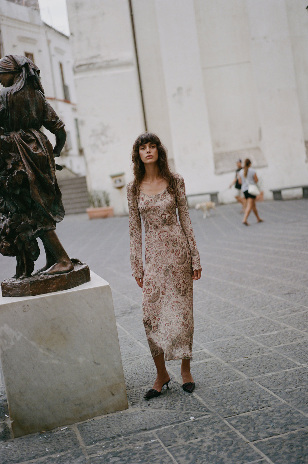 Josephine dress in stone paisley print with long sleeves, scoop neckline, and sheer finish standing in a stone plaza.