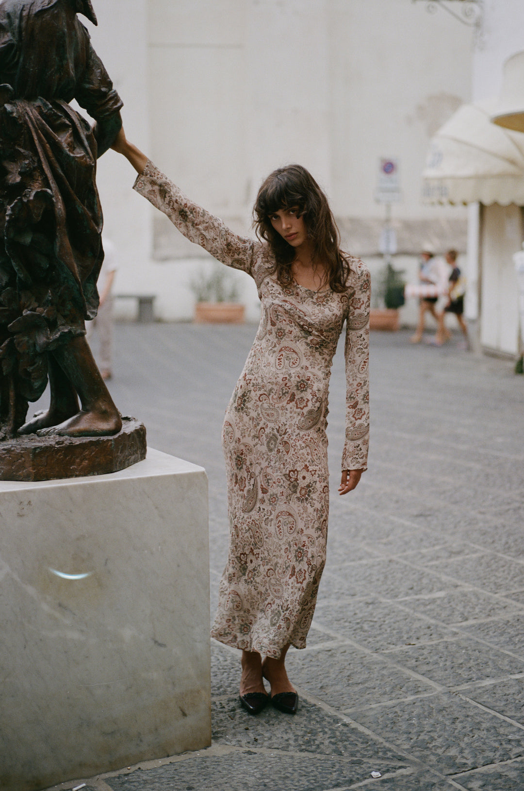 Josephine dress in stone paisley print with long sleeves, scoop neckline, and sheer finish standing by a statue.