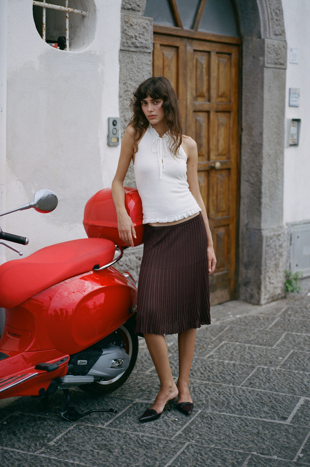 White knitted halter top with a self-tie neckline, button-front, and ruffle trim, styled with a brown pleated midi skirt.