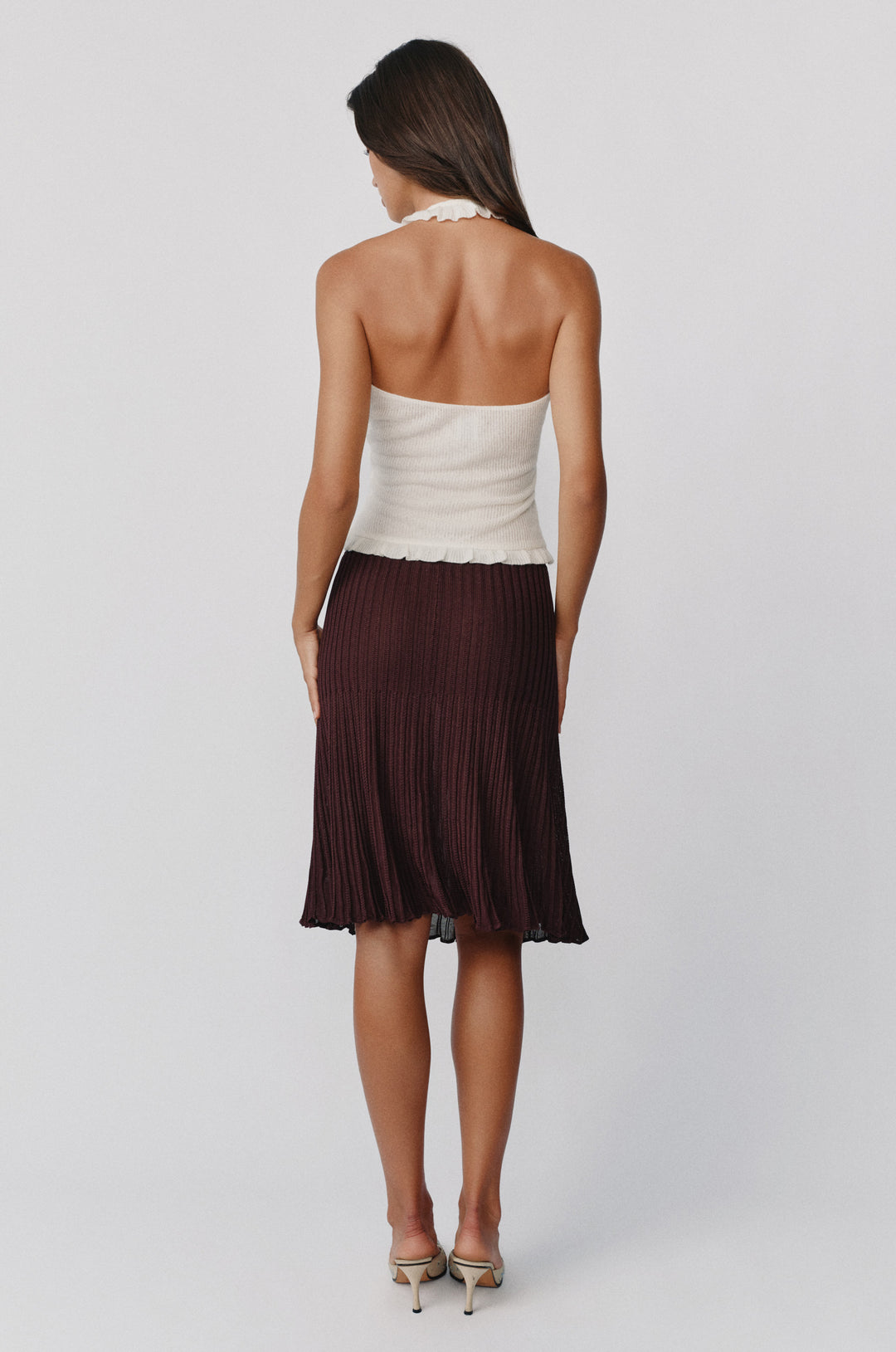 White knitted halter top with a self-tie neckline and ruffle hem, worn with a pleated dark brown midi skirt.