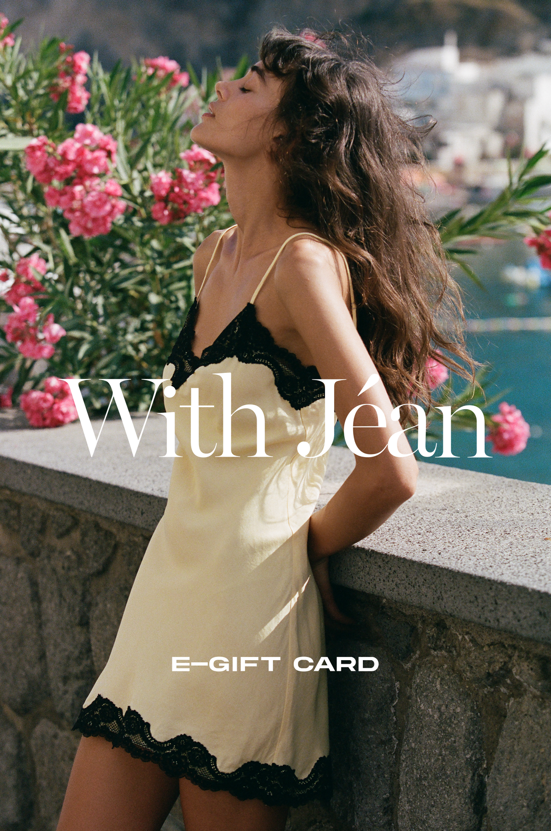 Woman in a pale yellow silk slip dress with black lace trim, eyes closed and face tilted toward the sun, leaning against a stone ledge with pink oleander blossoms and a coastal harbor in the background. "With Jéan" logo and "E-Gift Card" text overlaid.