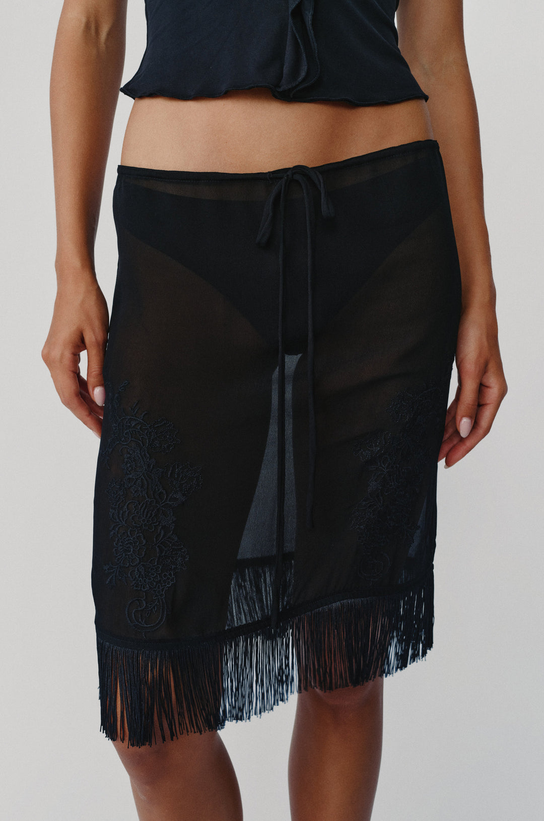 Black sheer low-rise Andie skirt with an adjustable drawstring waist, embroidered front detailing, and a fringe hemline.