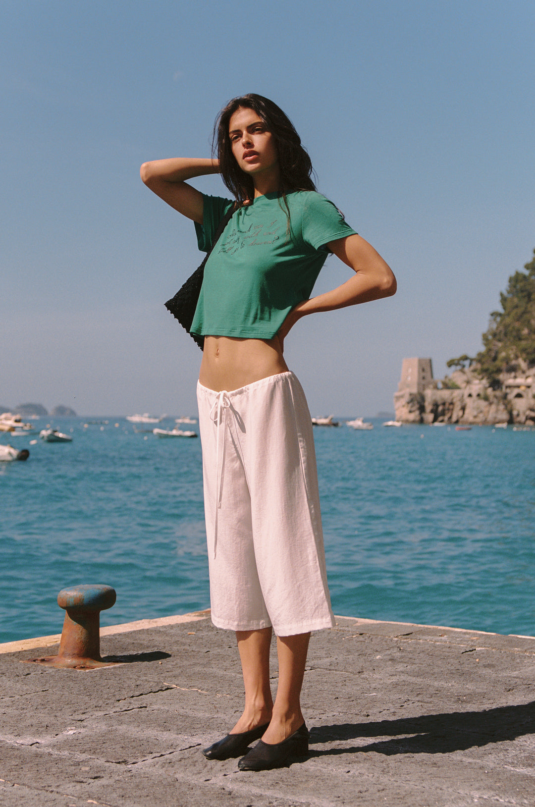 Woman in white Arden Capri Pant with adjustable tie waistband, green crop top, and black flats by the sea.