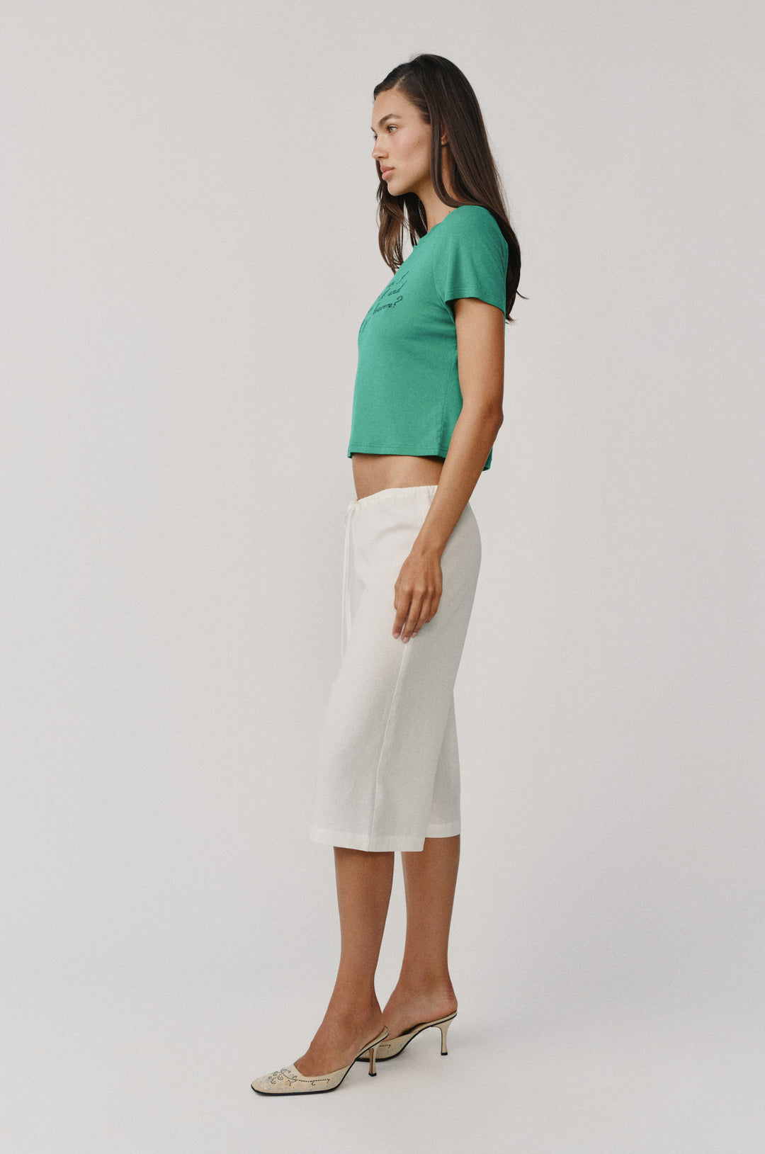 Woman in white Arden Capri Pant with adjustable tie waistband, green top, and embellished heels.