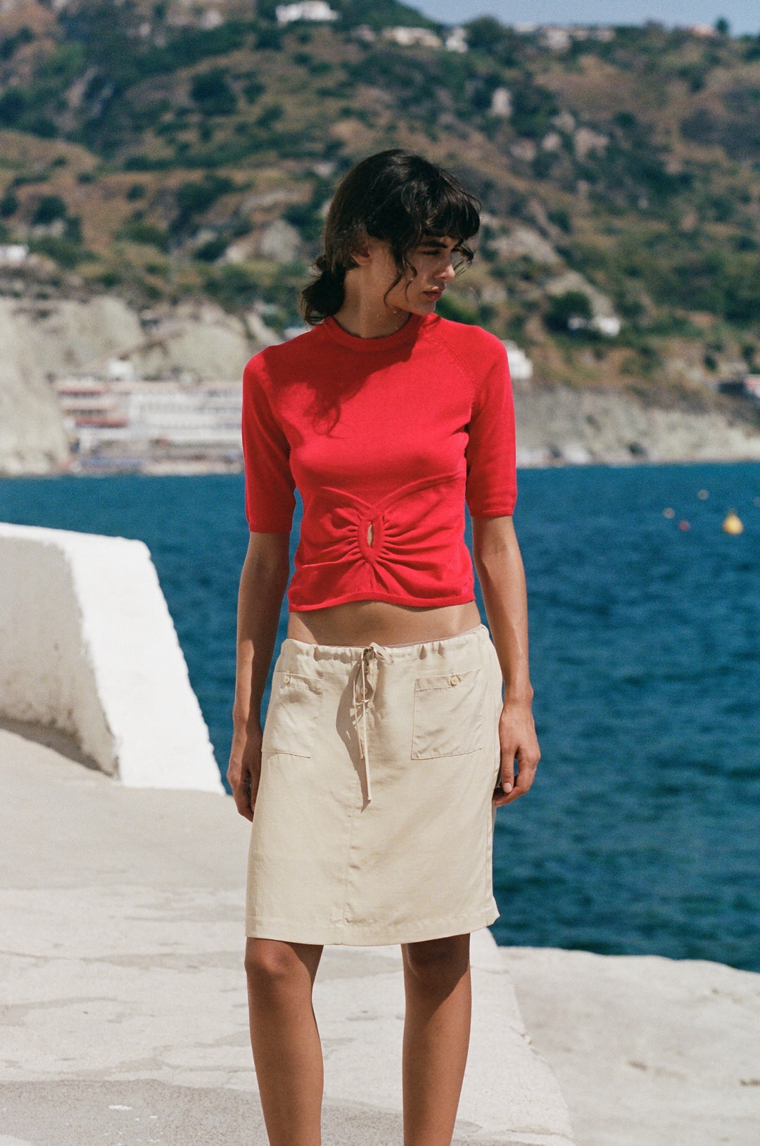 Red knit Avery top with half sleeves and a gathered front keyhole cut-out, worn with a beige skirt by the sea.