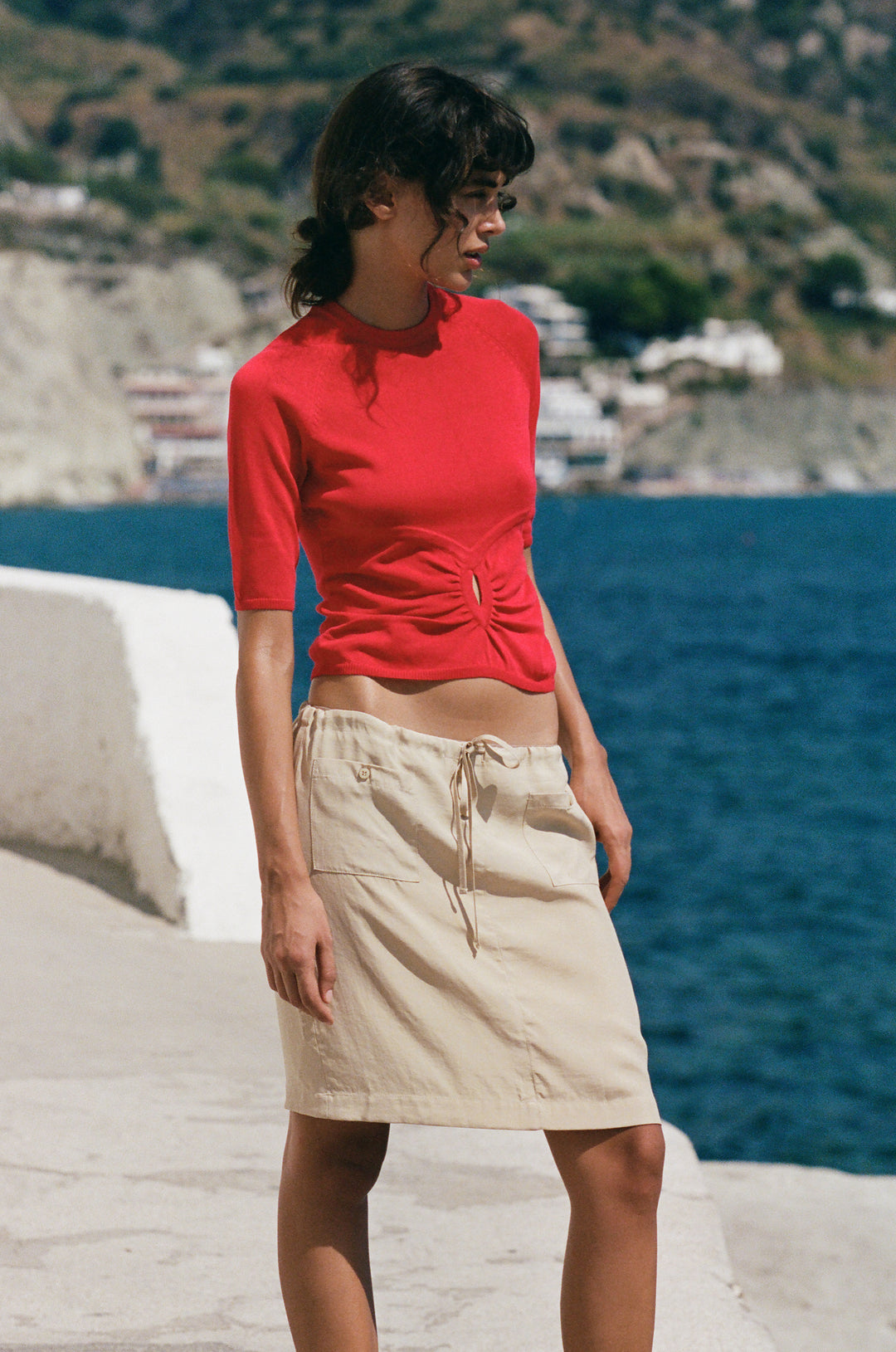 Red knit Avery top with half sleeves and a gathered front keyhole cut-out, paired with a beige skirt by the sea.