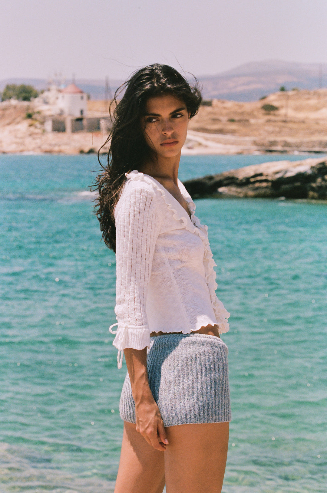 White knitted Bianca top with 3/4 sleeves, ruffle-trim neckline, and tie-detail cuffs worn by the ocean.