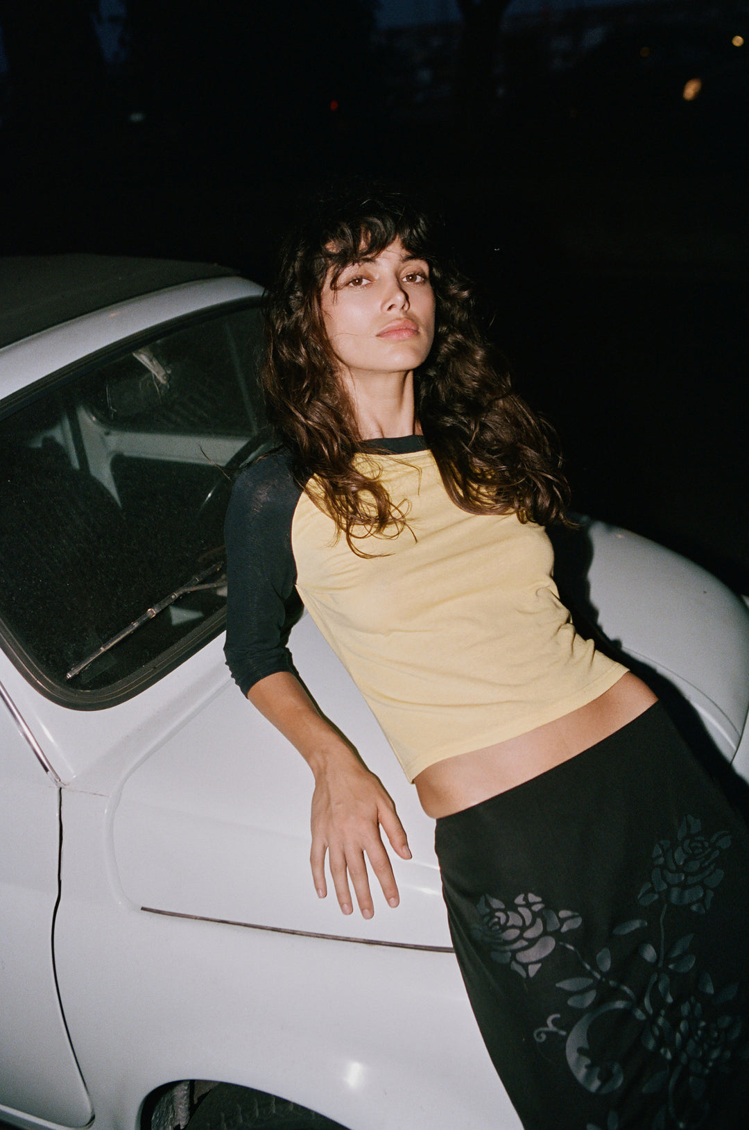 Woman wearing a yellow and midnight navy raglan tee with a round neckline, leaning against a white car at night.