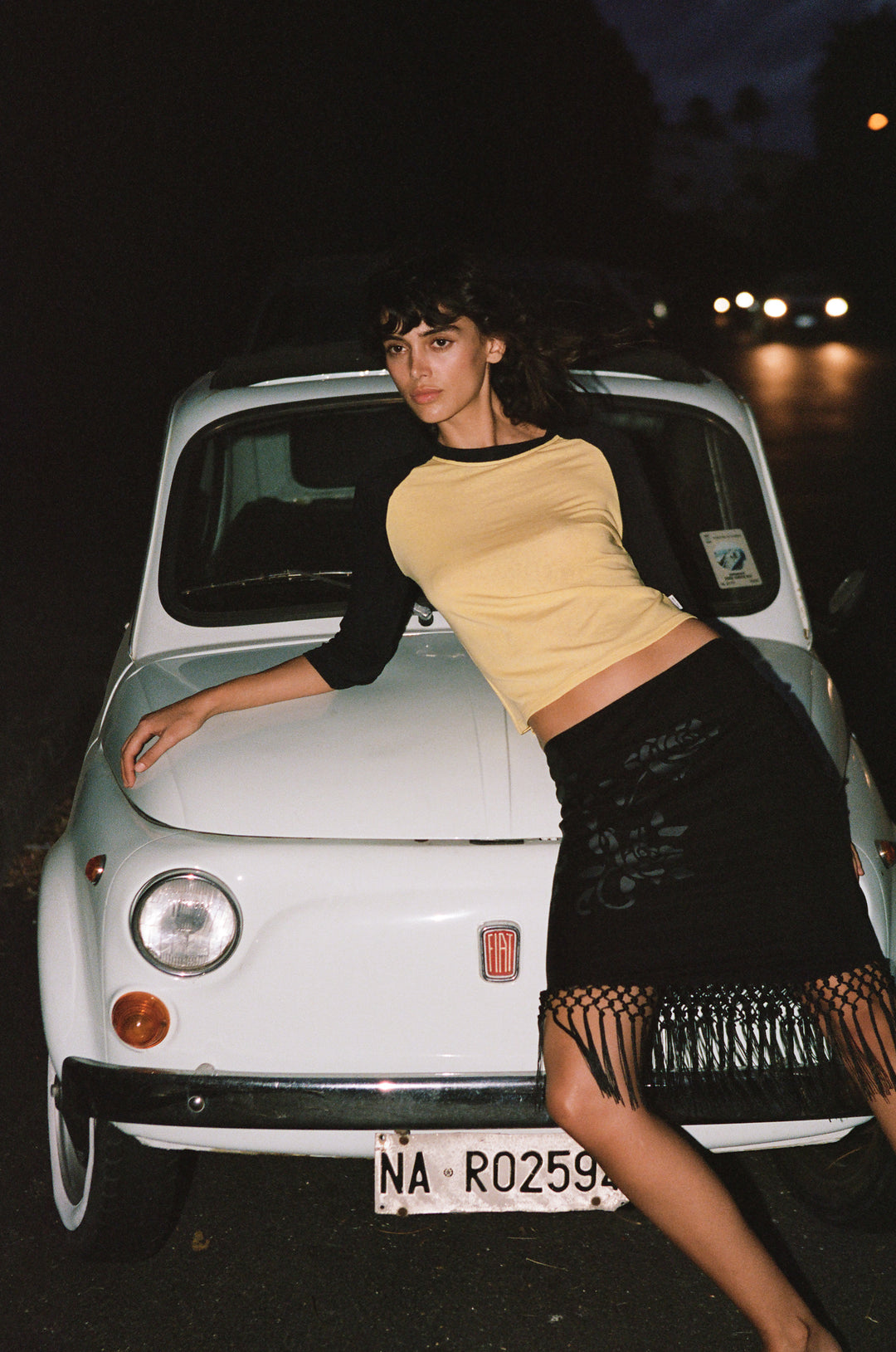 Model wearing a yellow and midnight navy raglan tee with a round neckline, leaning against a vintage white Fiat car at night.