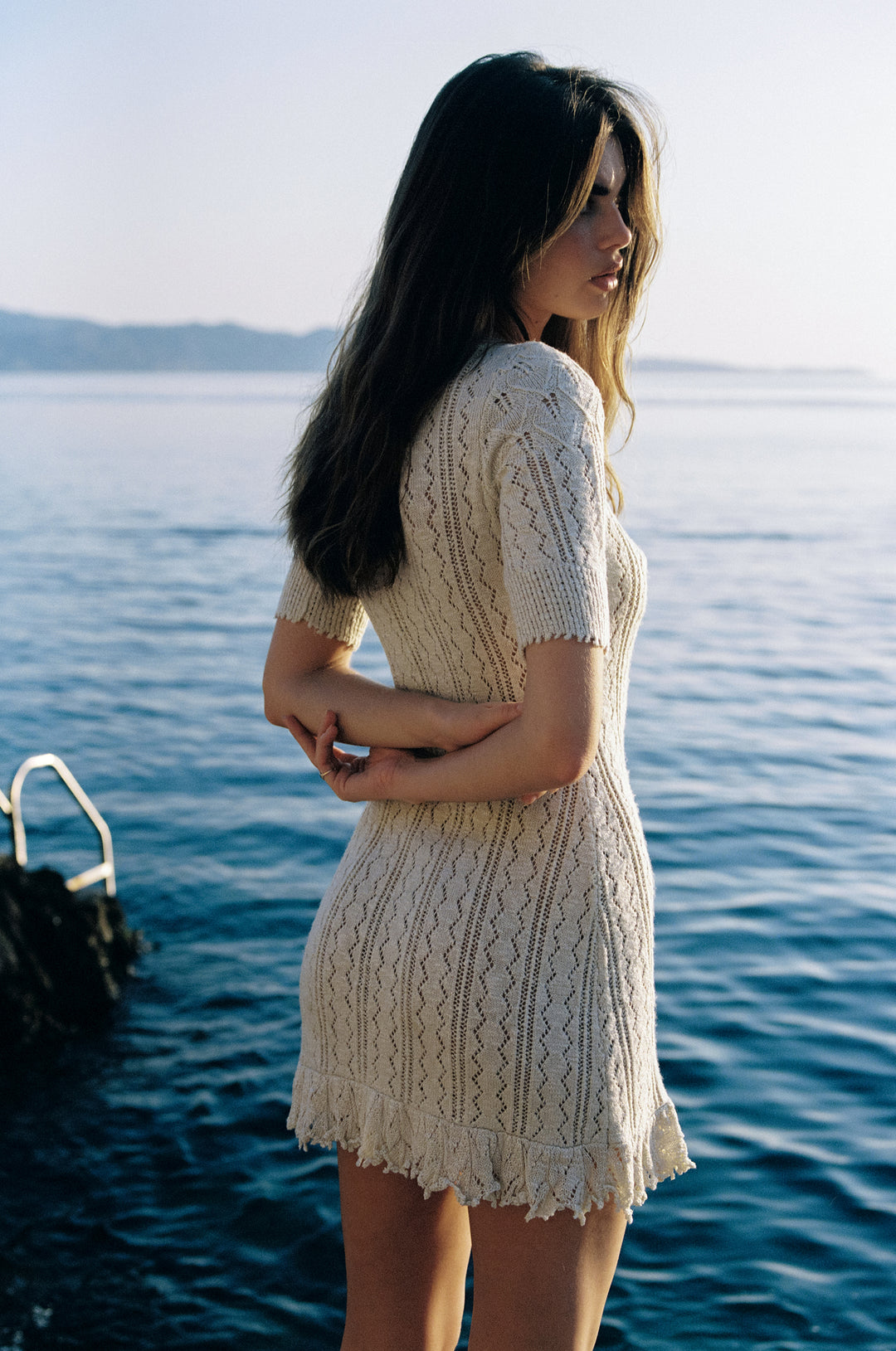 Woman wearing a pearl-colored mini dress with openwork knit construction, ribbed scoop neckline, and textured hem by the sea.