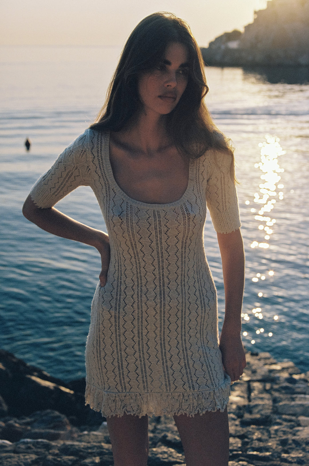 Pearl mini dress with openwork knit, ribbed scoop neckline, and textured hem worn by the ocean at sunset.