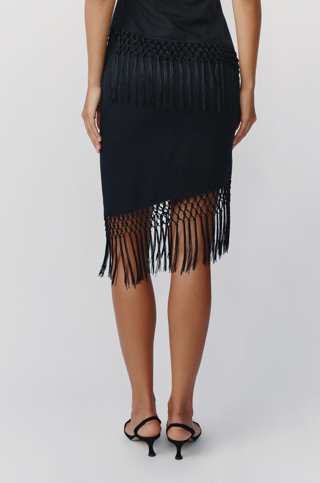 Black low-rise Daria skirt with an asymmetrical hem and decorative macrame fringe detailing.