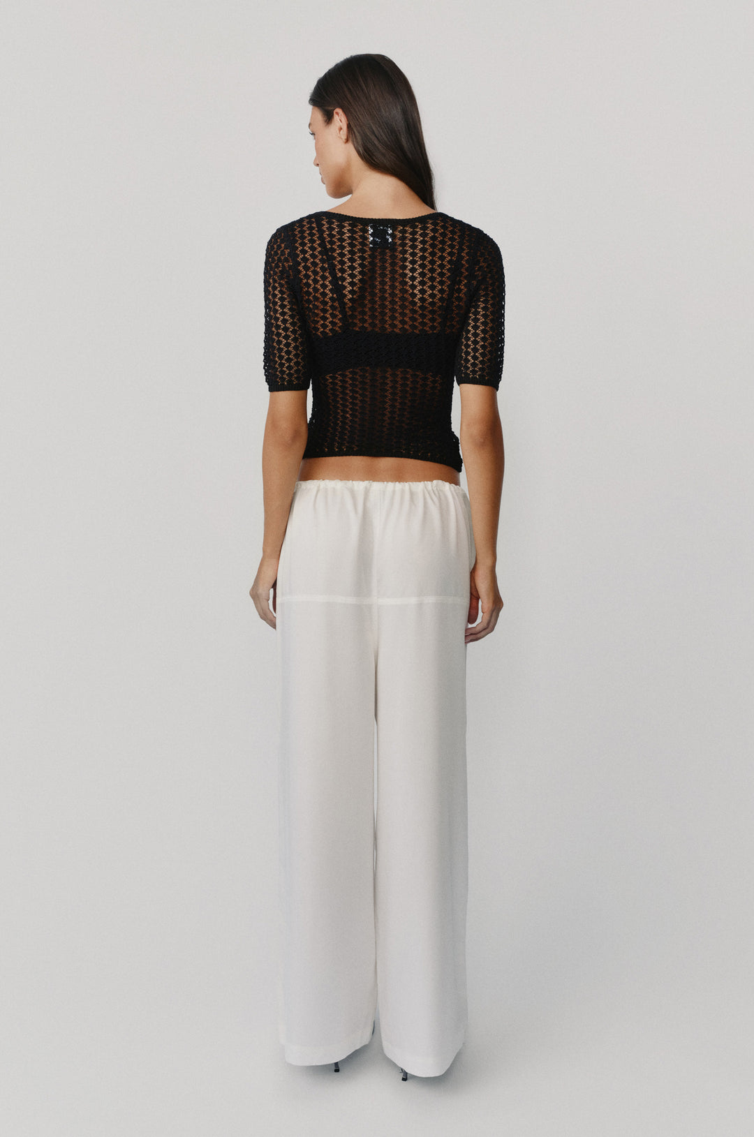 Back view of a black knitted Delilah top with half sleeves and a textured open-knit pattern worn with white trousers.