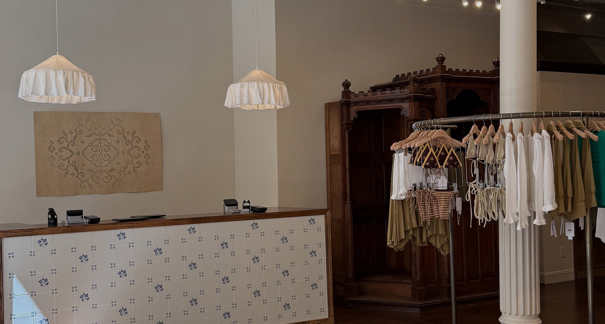 A boutique interior with a patterned counter, two white pendant lights, a dark wooden cabinet, and clothing racks holding white and olive garments.