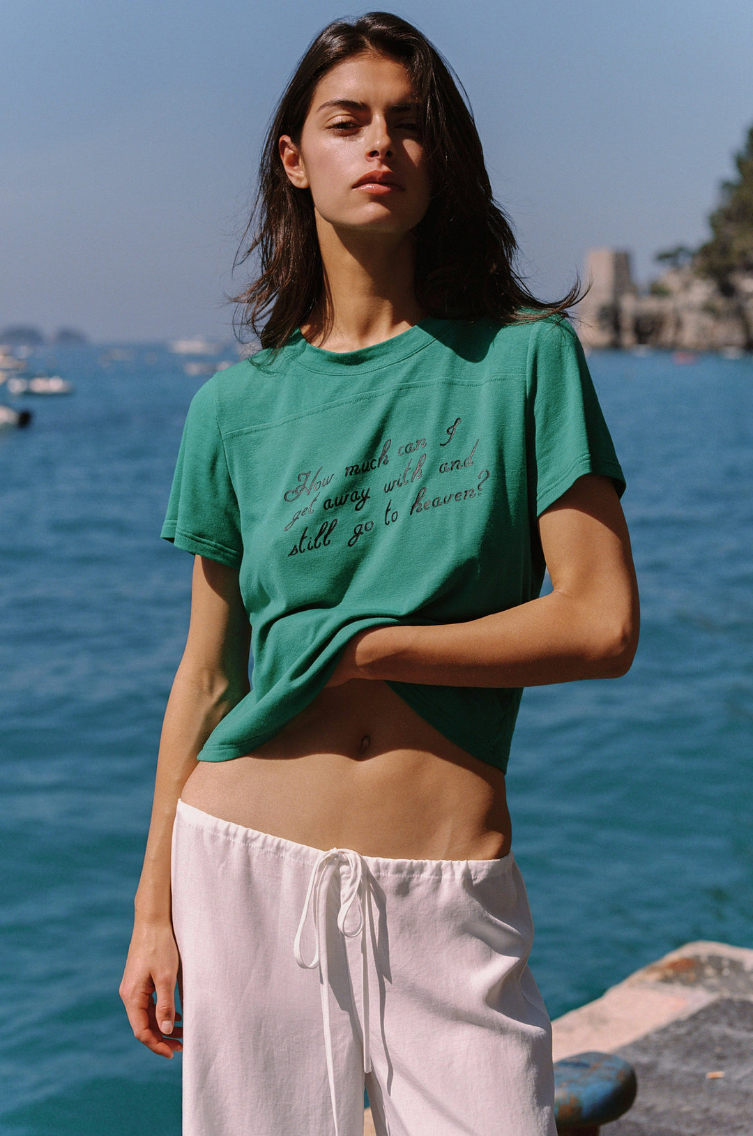 Woman wearing an emerald green Ellie Tee with "How much can I get away with and still go to heaven?" text, paired with white drawstring pants by the sea.