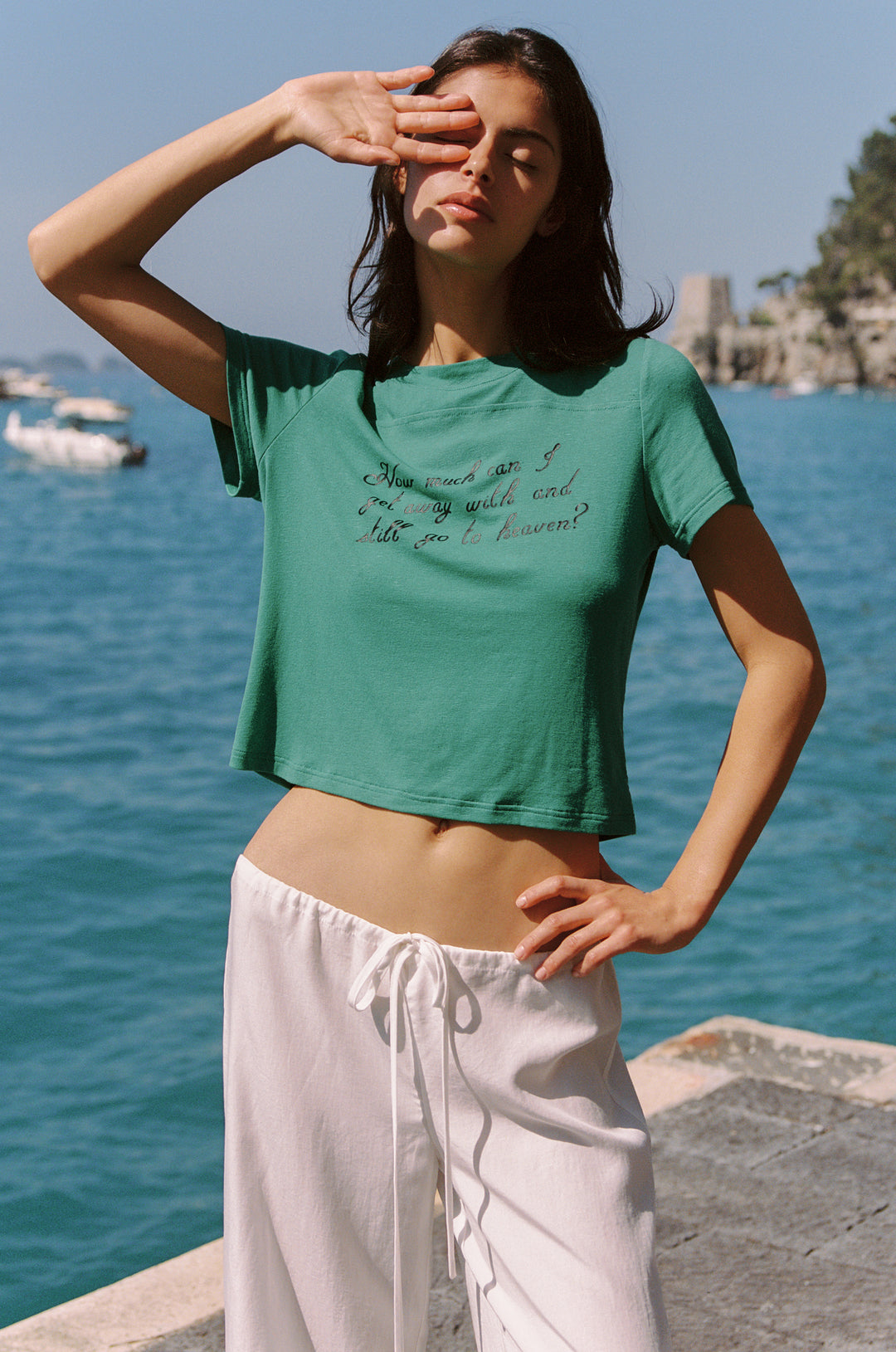 Woman in emerald Ellie Tee with "How can I go wrong with you and still go to Heaven" text, white pants, by the sea.