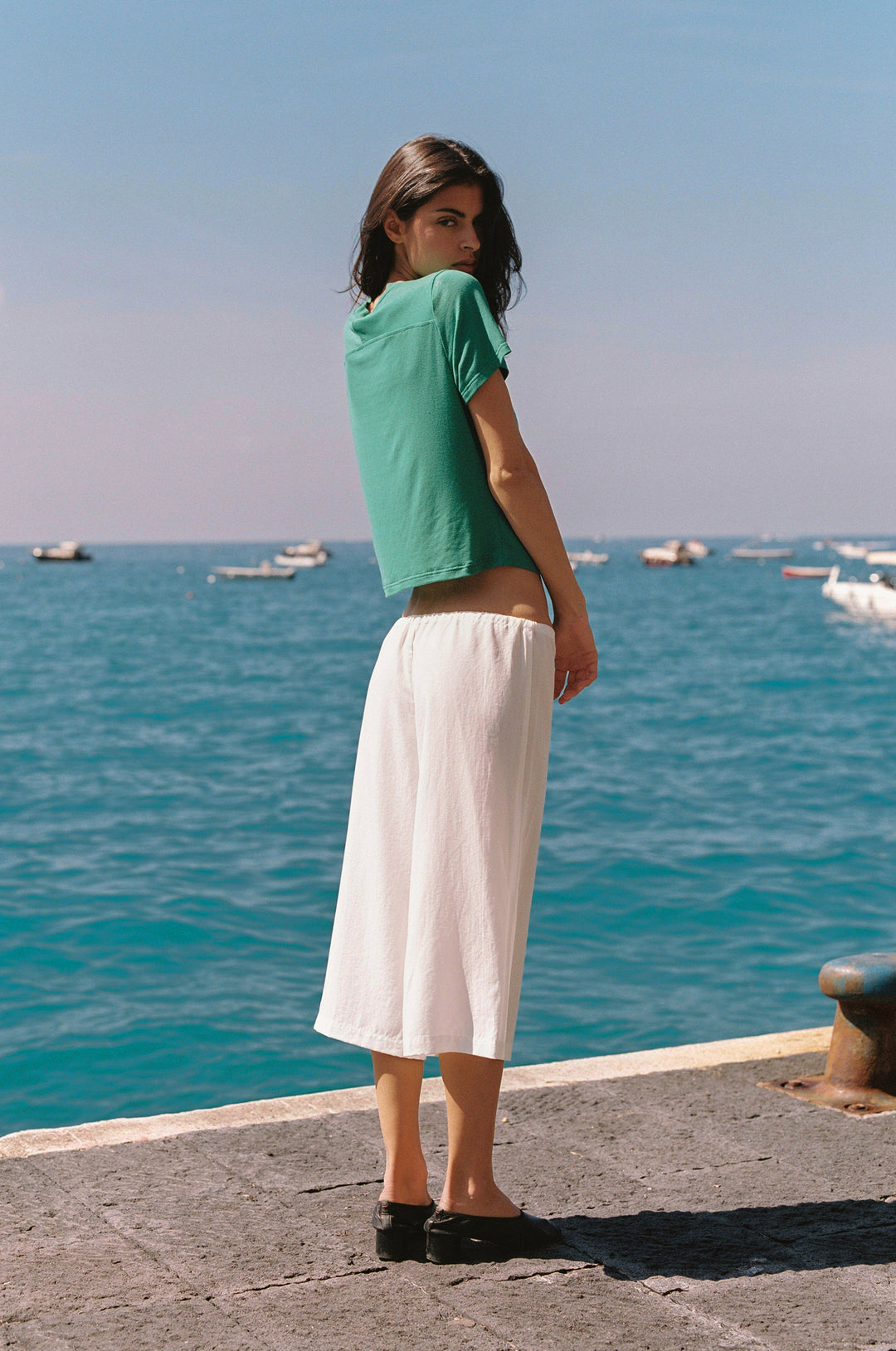 Woman in emerald green Ellie Tee with short sleeves and round neckline, paired with white wide-leg pants, standing by the ocean.