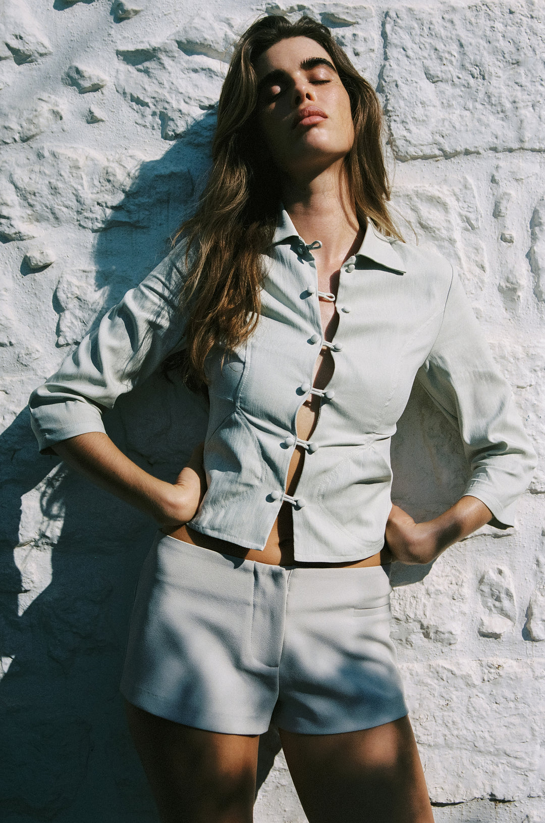 Low-rise dusty sage mini Hudson shorts paired with a matching button-down top against a white stone wall.