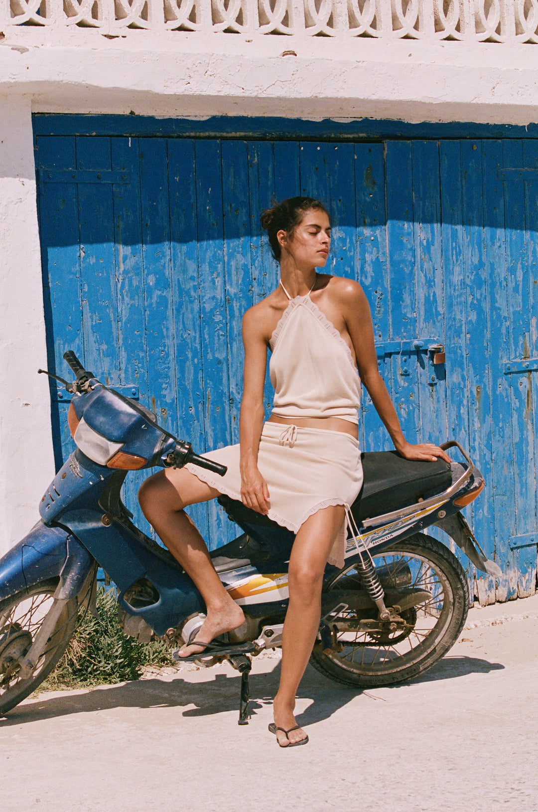 Beige knitted halter top with scalloped contrast trim, worn with a matching skirt while sitting on a blue motorbike.