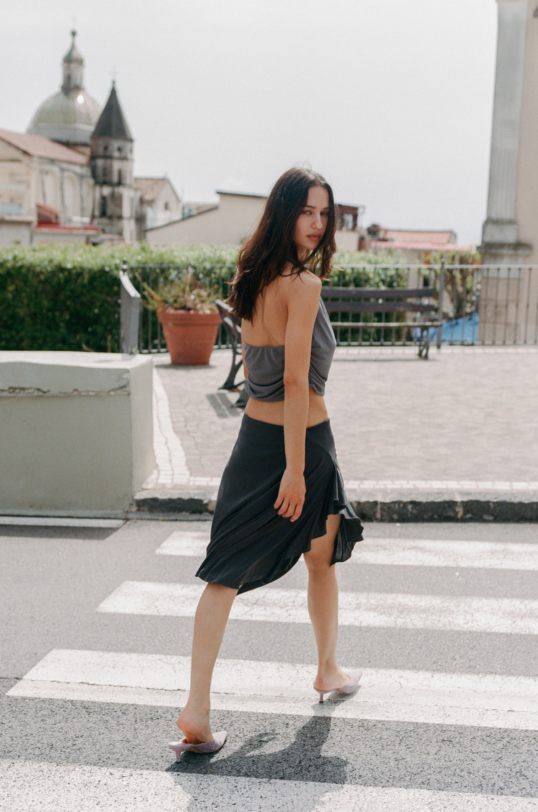 Woman in a slate Jolene halter top with ruched sides and a dark skirt, walking on a crosswalk.