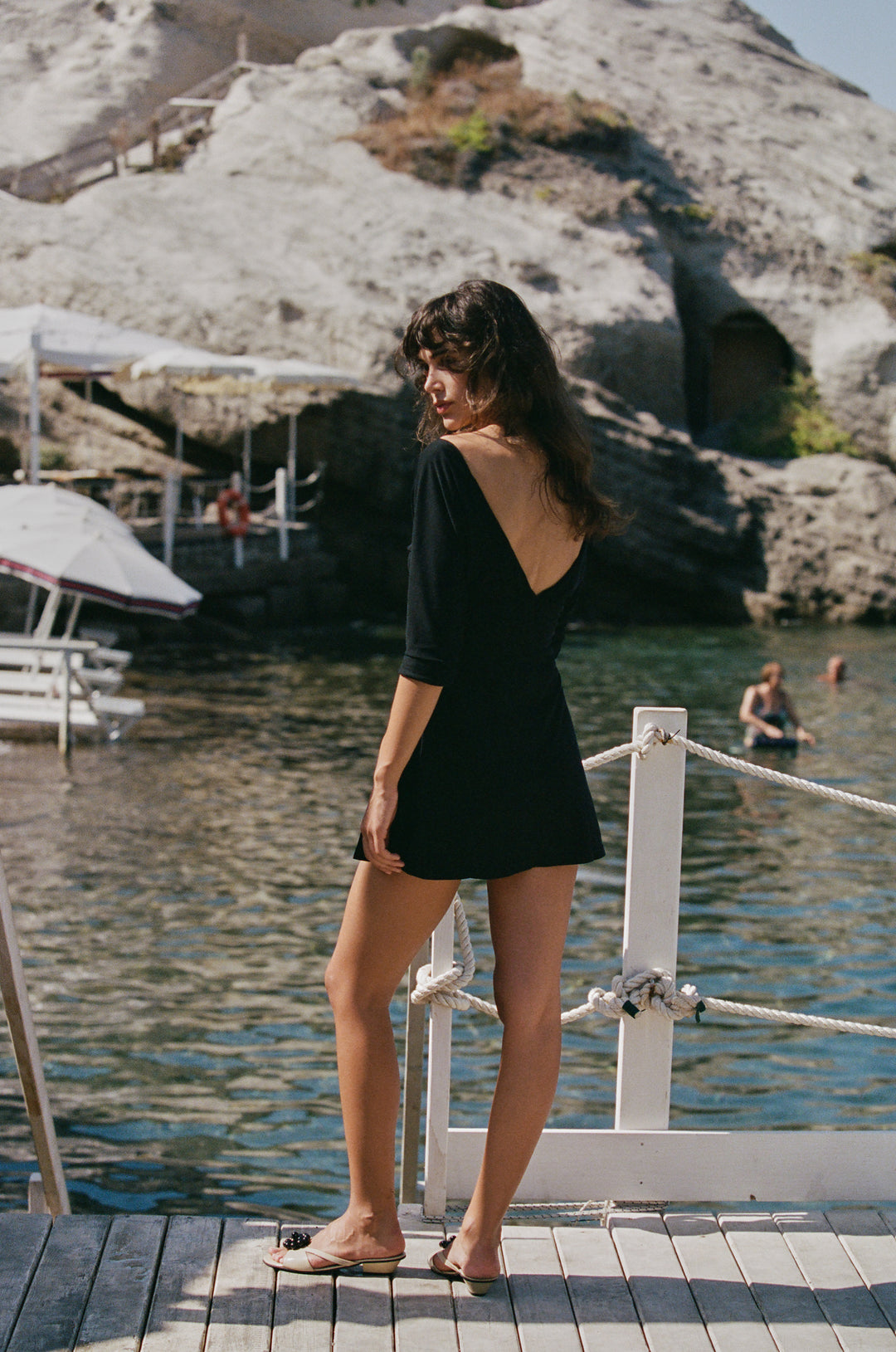Black mini Lana dress with a V-neckline and half sleeves, worn on a wooden dock by the sea.