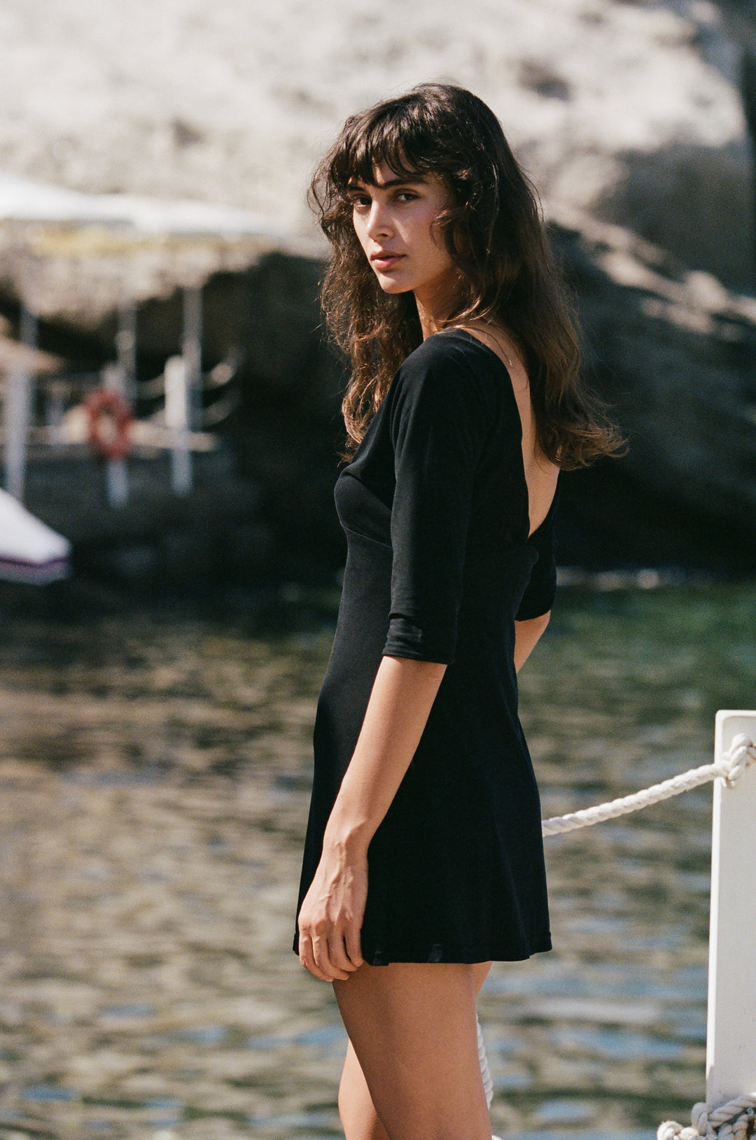 Model wearing a black mini Lana dress with half sleeves and a V-neckline, standing by the water.