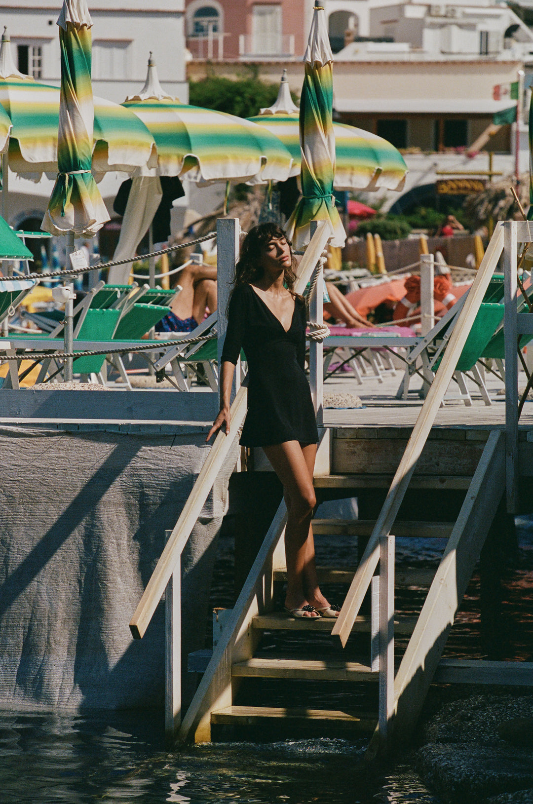 Woman wearing a black mini Lana dress with a V-neckline and half sleeves, standing on a wooden pier by the beach.