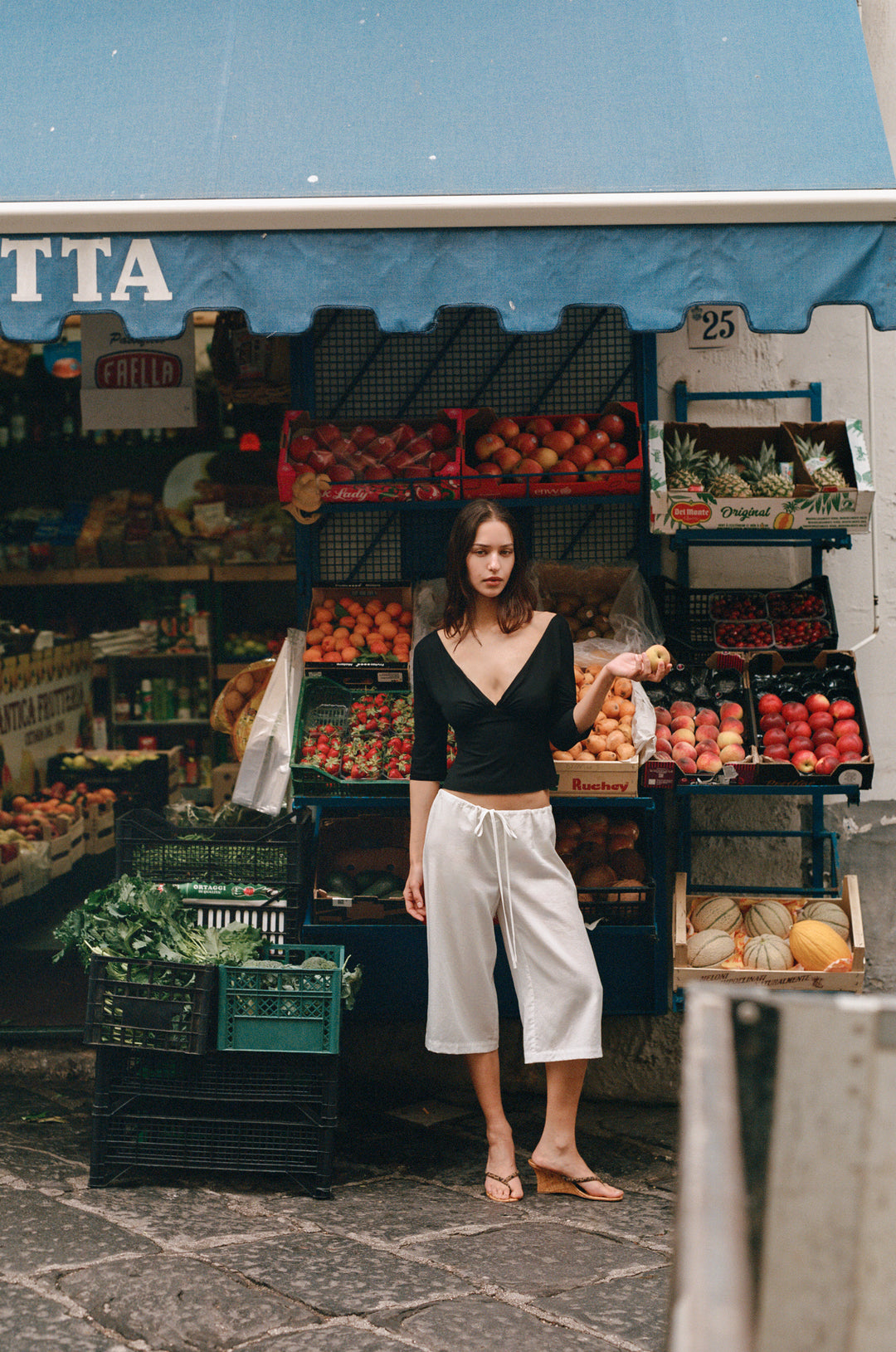Woman in a black Lana Top with V-neck, half sleeves, and gathered bust, standing in front of a fruit stand.
