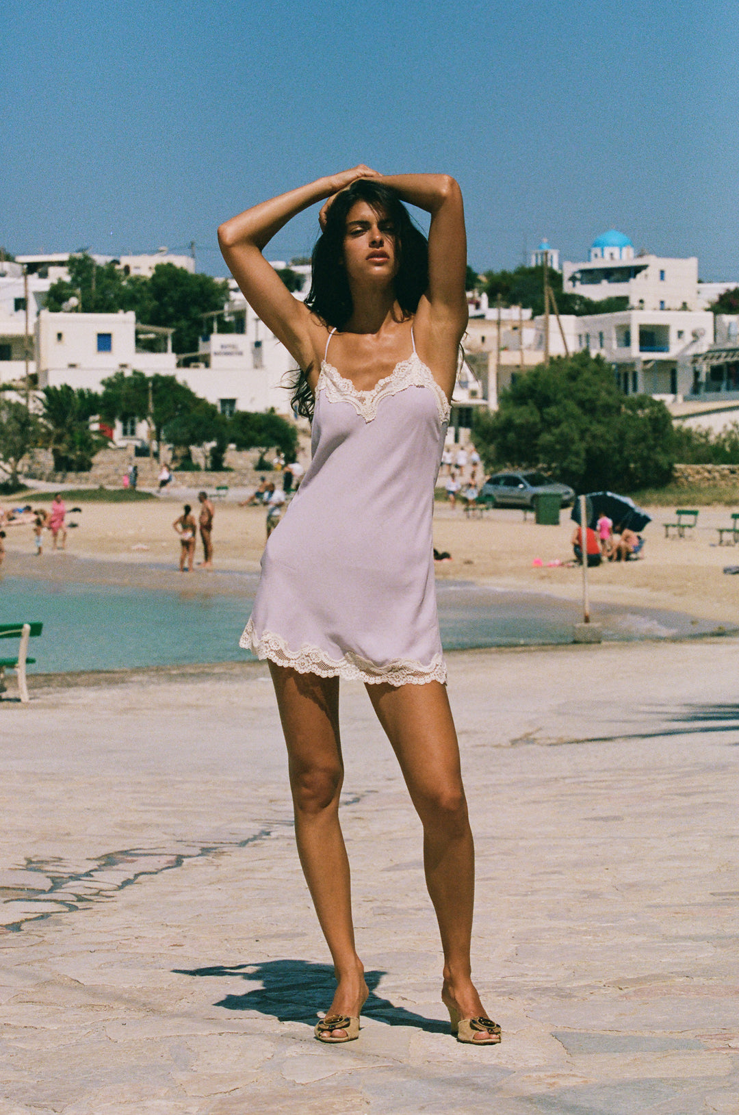 Lilac mini slip dress with adjustable straps and delicate cream lace trim at the neckline and hem on a beach.