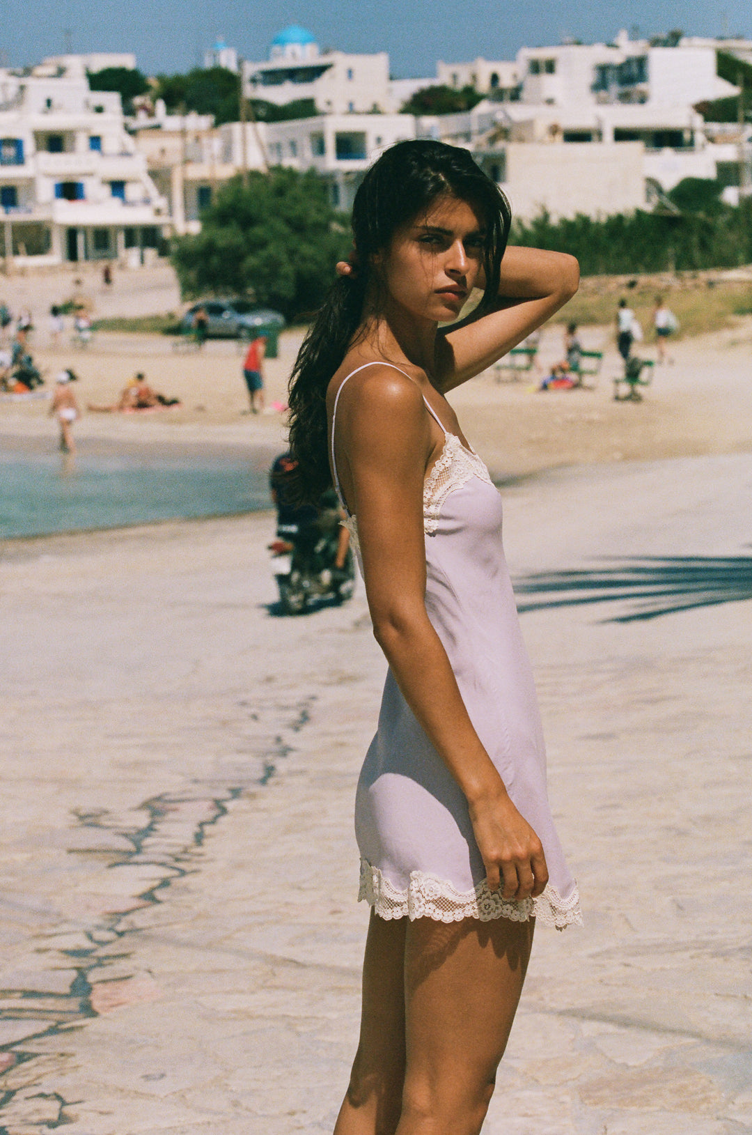 Lilac mini slip dress with adjustable straps and delicate cream lace trim at the neckline and hem on a sunny beach.