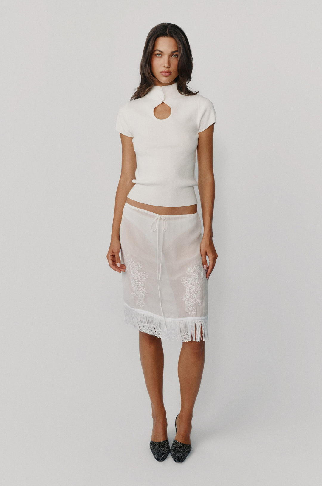 White knitted Luci top with a high neck, button closure, and front keyhole cutout, paired with a sheer fringed skirt.