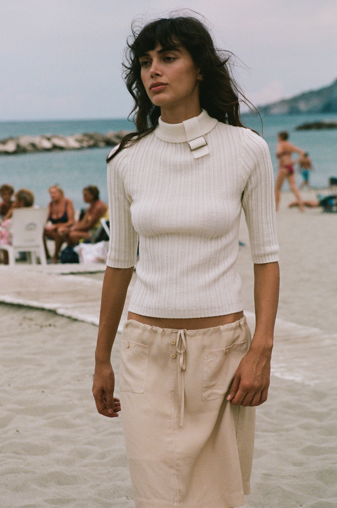 Off-white fine wool-knit top with a high neckline, adjustable buckle detail, and half sleeves worn on a beach.
