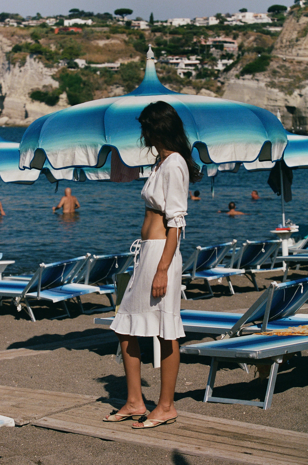 White low-rise Maisie skirt with an adjustable drawstring waist and asymmetrical ruffle hem, worn on a beach.