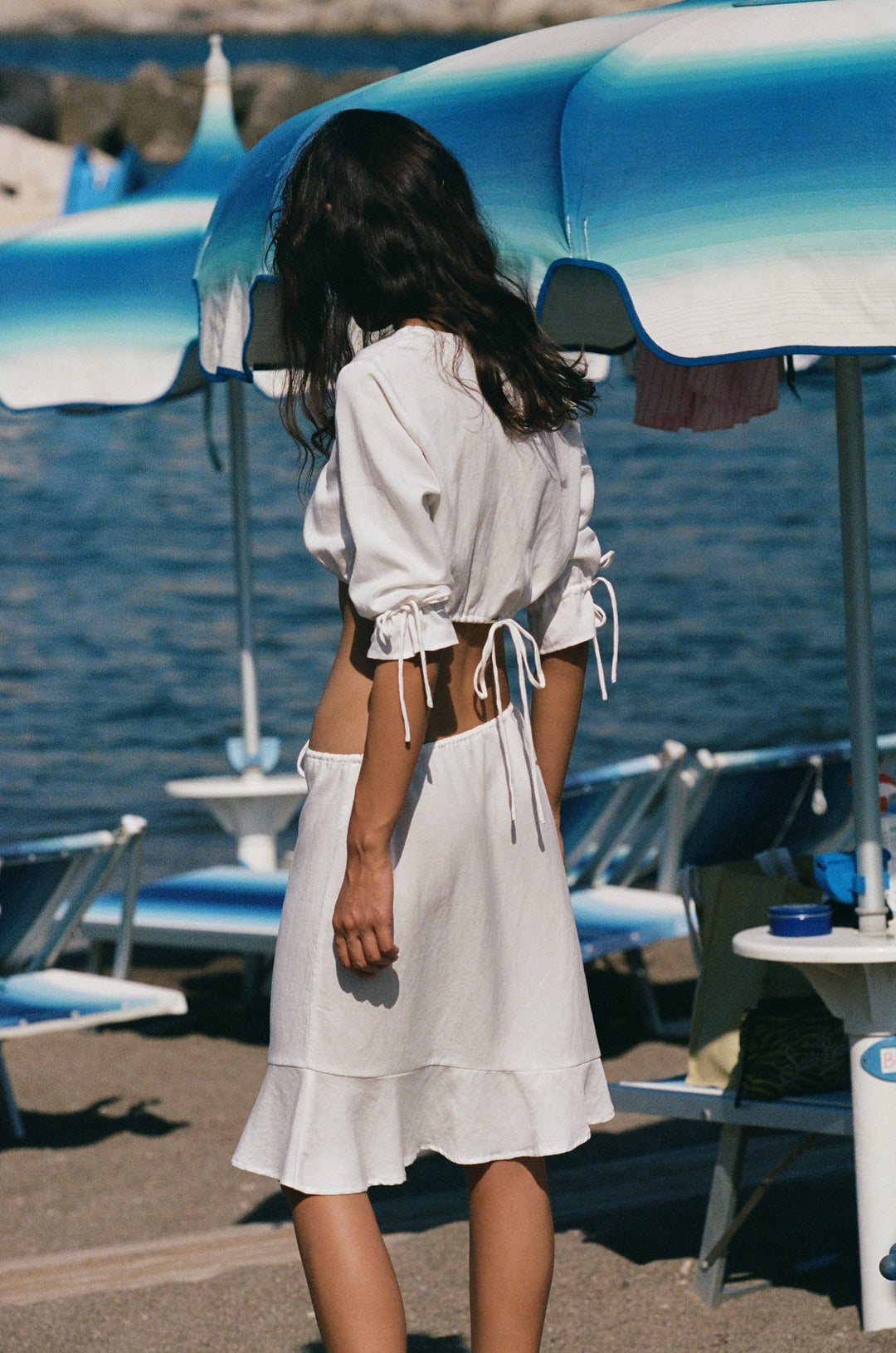White low-rise Maisie skirt with an adjustable drawstring waist and asymmetrical ruffle hem, worn on a beach.