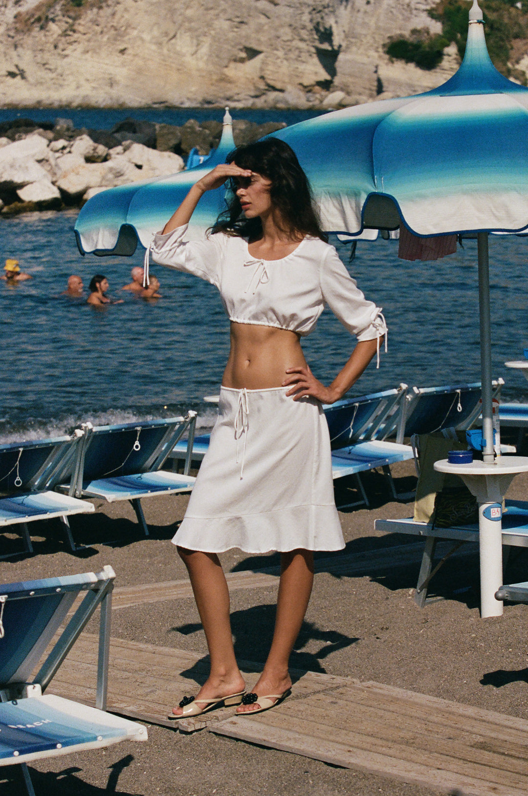 White low-rise Maisie skirt with an adjustable drawstring waist and asymmetrical ruffle hem on a beach.
