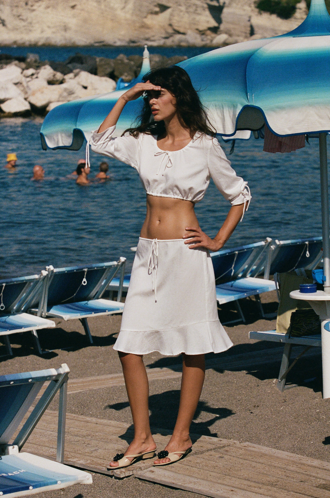 White cropped Maisie top with scoop neck, tie details, and 3/4 ruffle sleeves worn on a beach.
