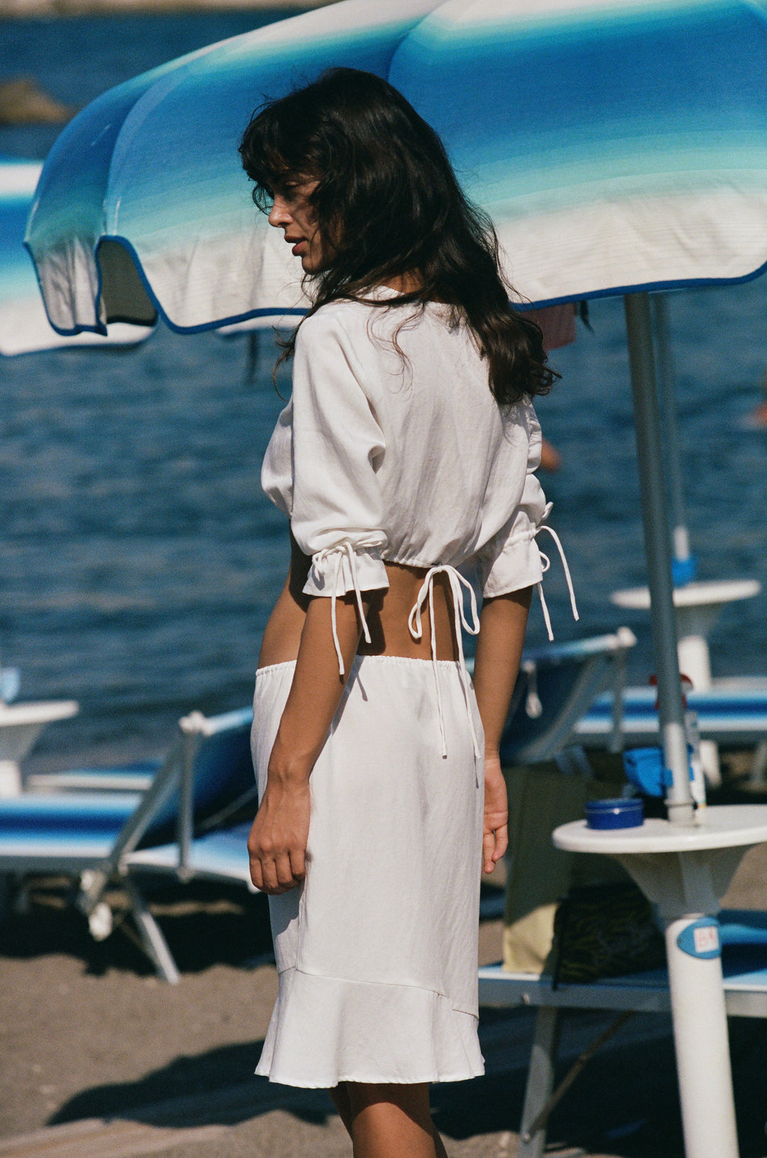 White cropped Maisie top with 3/4 sleeves, ruffle cuffs, and adjustable drawstring hem worn at the beach.