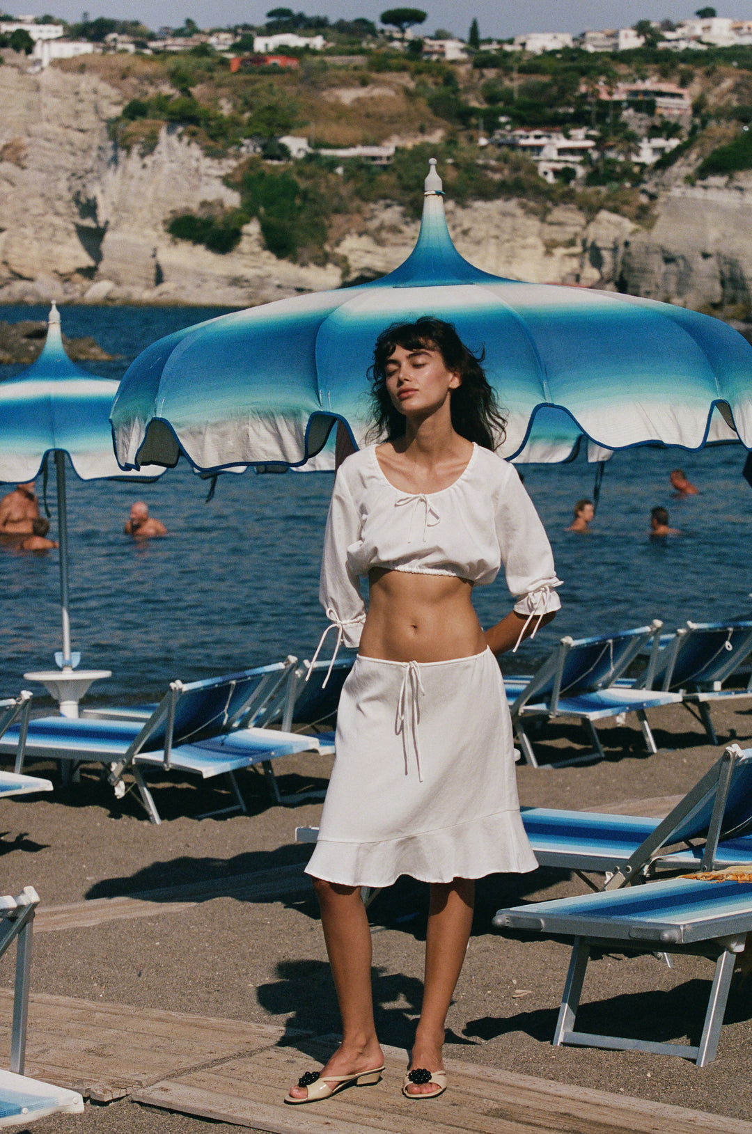 White cropped Maisie top with scoop neckline, 3/4 ruffle sleeves, and drawstring hem worn on a sunny beach.