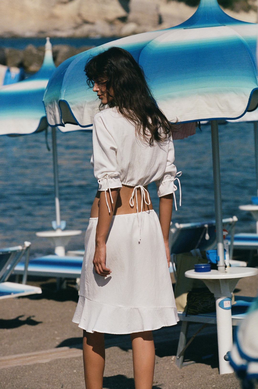 White cropped Maisie top with 3/4 ruffle sleeves, tie details, and adjustable drawstring hem worn on a beach.