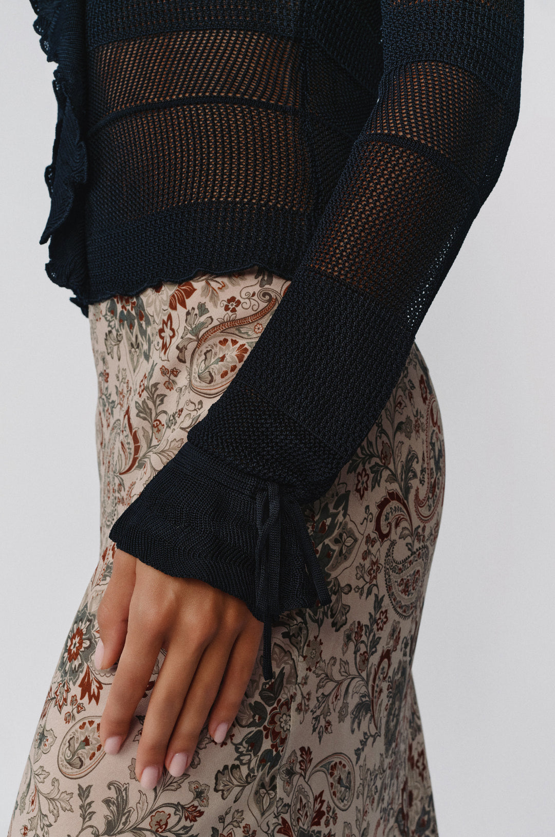 Black knitted long-sleeve top with ruffle trim and tie-detail cuffs worn over a patterned skirt.