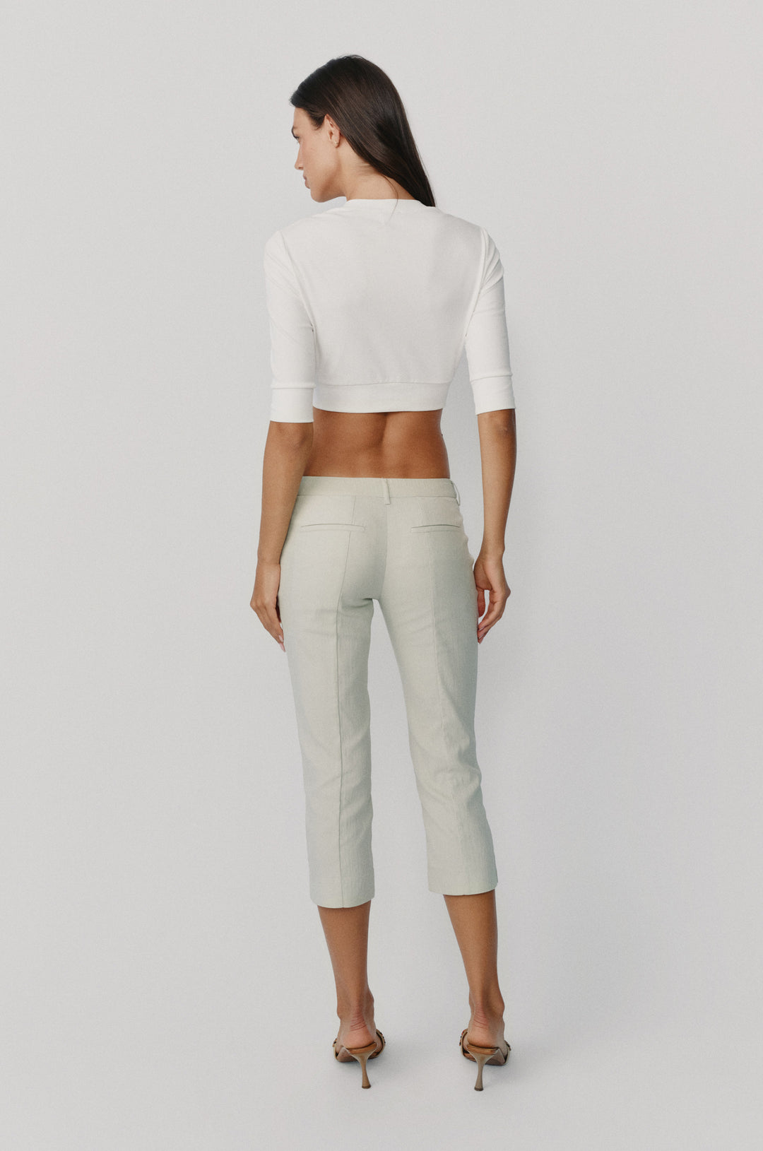 Back view of a woman wearing a white cropped half-sleeve top with a fitted underbust band and light-colored capri trousers.