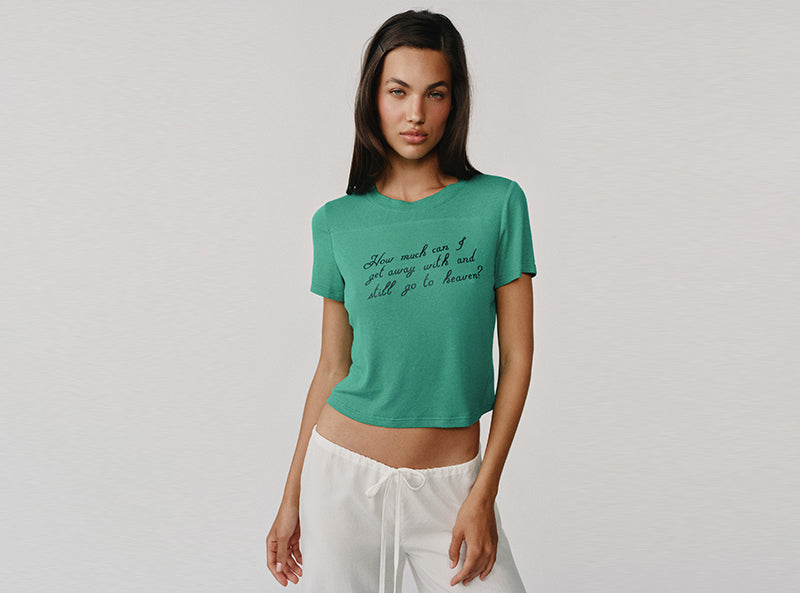 A woman modeling a green short-sleeve crop top with black cursive text on the front, paired with white drawstring pants, against a light gray background.