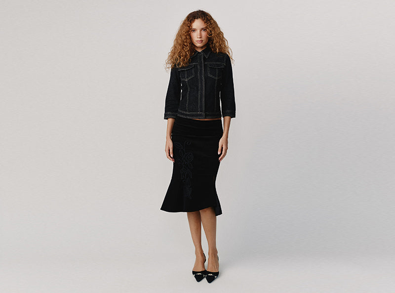 A model with curly light brown hair stands against a light gray studio background wearing a black denim jacket and a black velvet midi skirt with a subtle tonal pattern.