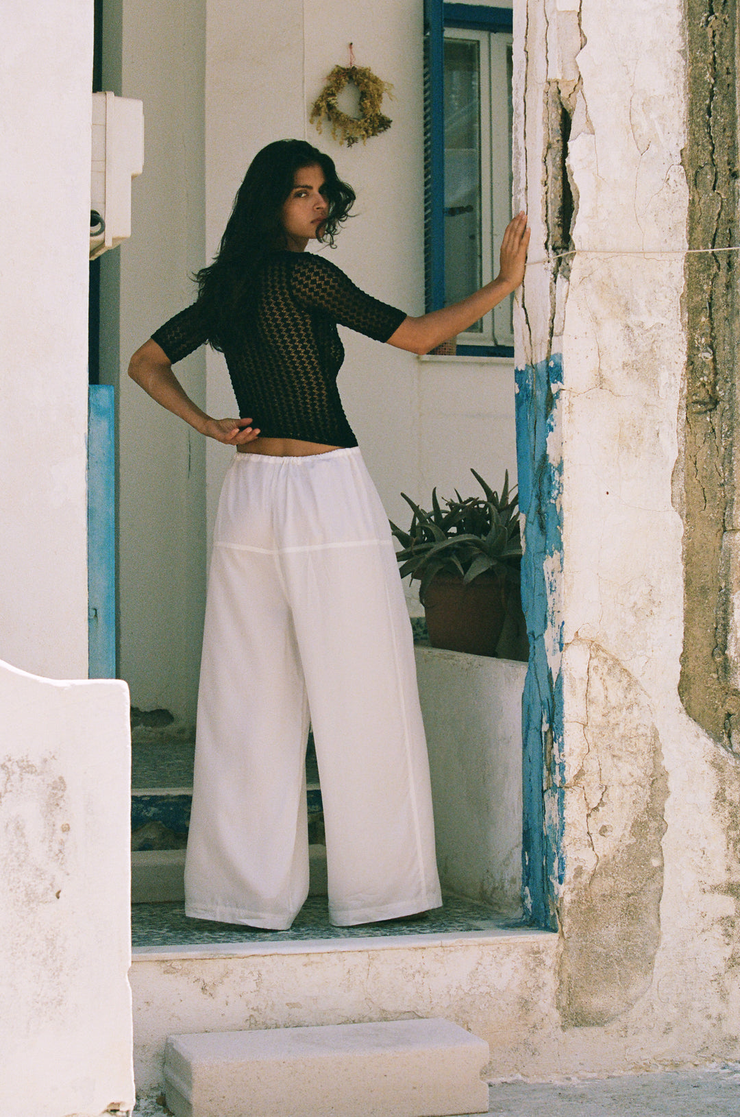 Woman wearing loose-fit white Norah pants with a self-fabricated tie waistband, standing in a doorway.