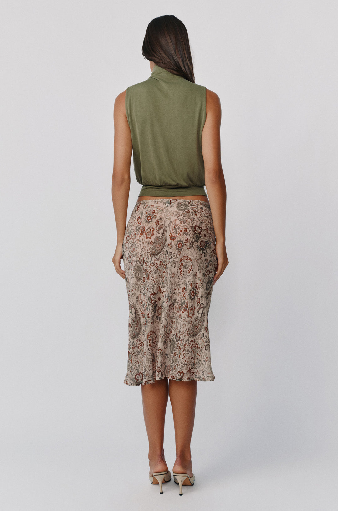 Back view of a sleeveless army green Nova top with a high neck and fitted hem, paired with a patterned midi skirt.