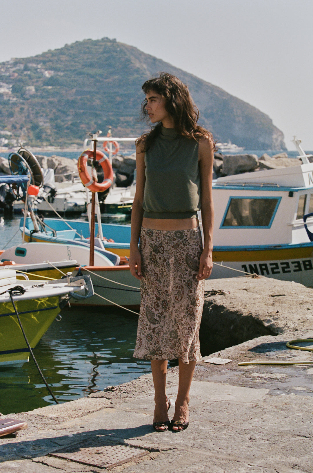 Woman wearing a sheer, low-rise Olea midi skirt in a stone paisley print, styled with a sleeveless olive green top.
