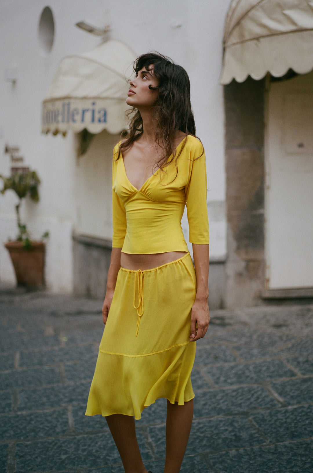 Golden yellow sheer Pali skirt with an adjustable drawstring waist and flowy drop seam hemline worn on a cobblestone street.