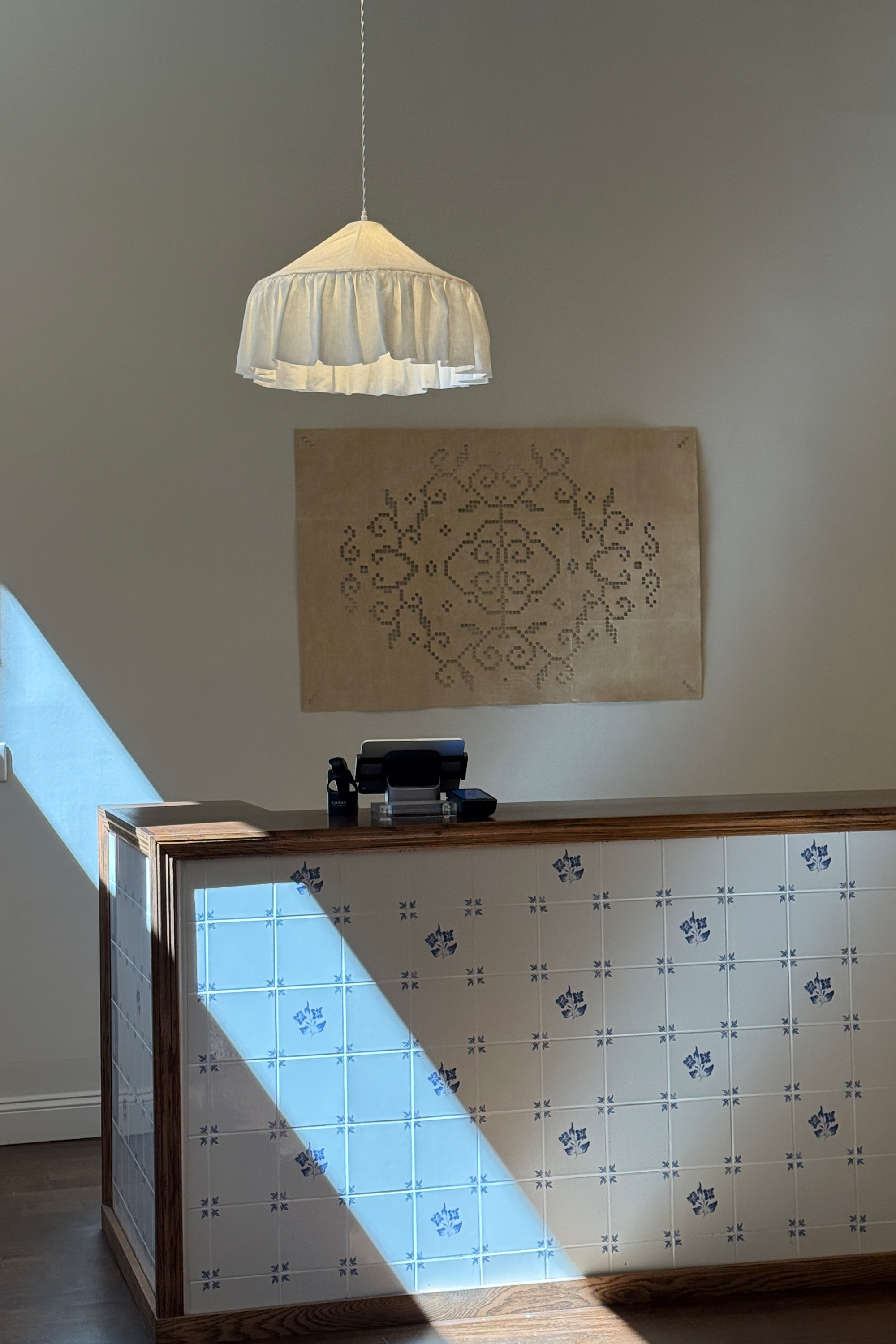 A modern interior scene featuring a wooden reception desk with blue and white patterned tiles, a hanging pleated white pendant light, and a beige wall hanging with a dark floral design. Sunlight casts a strong diagonal shadow on the desk and wall.
